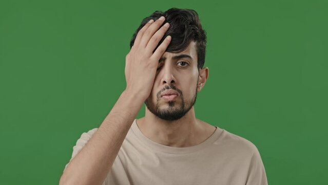 Male portrait of arabic indian disappointed guy upset stressful man stand in green studio cover face with hand outrage puts palm on head shame lost problem feeling exhaustion shows unpleasant emotion