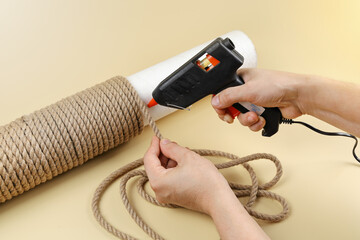 Hands hold electric hot glue gun to make rope around column for scratching post for cats