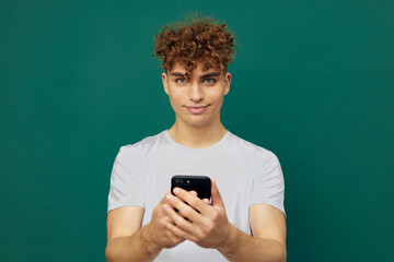 a funny, joyful man with curly hair stands against a green background and holds his smartphone with both hands, smiling pleasantly and looking at the camera