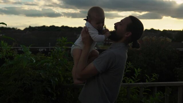 Happy Father holding adorable baby boy embracing kissing. Smiling Dad with son playing outside Garden. Family silhouette at sunset. Daddy fly up child as airplane having fun. Parent enjoying with kid