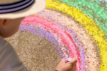 A happy child draws a rainbow with chalk on the asphalt outdoors. Portrait of a boy drawing with...