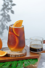 cold Iced black coffee mixed with orange juice and orange slices in glass on wooden plate in white background with nature leaves silhouette shadow. two tones layer fresh summer drink in the morning
