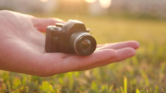 A small toy camera in the palm of a girl against the backdrop of a sunset and flowers on the street. 