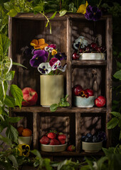 Summer still life with various ripe fruits and berries in wooden box and colorfull flowers surrounded by green plants.