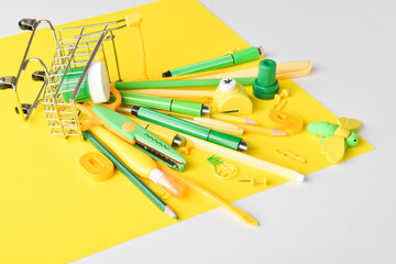 Various yellow and green stationery items are lying in a supermarket cart on a yellow background.