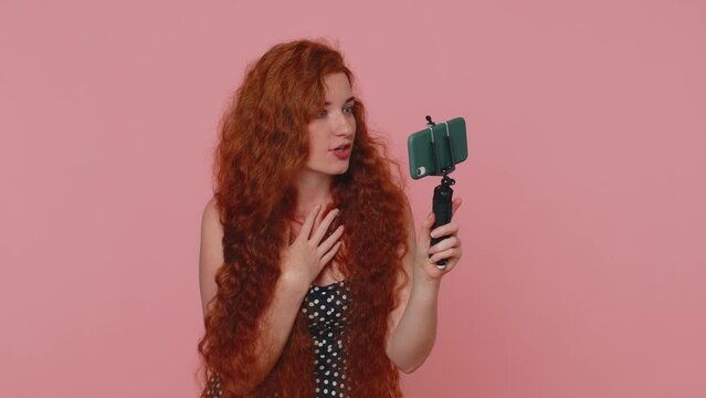 Wow reaction. Impressed redhead woman blogger taking selfie on mobile phone selfie stick, communicating video call online with subscribers. Young girl ginger isolated alone on pink studio background