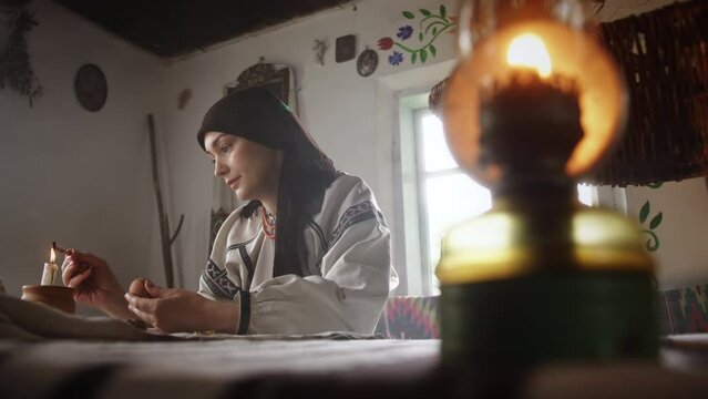 Young woman makes painted Easter egg at table lit by candle. Woman lit by candle makes Easter painted eggs at table in ethno museum house. Mother wearing Ukrainian national outfit looks concerned