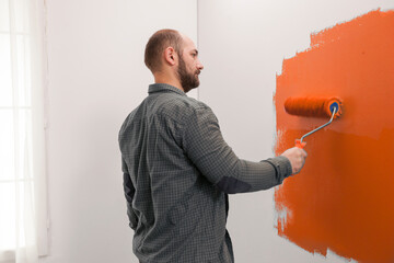 Man renovating apartment with orange paint, using new color on living room wall. Redecorating house with painting tools and paintbrush to change design, home decoration improvement.