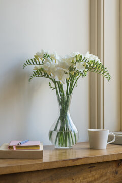 Vertical view of white freesia flowers in glass vase on oak table with cup and books next to window (selective focus)