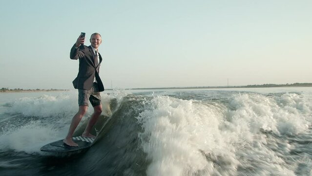A surfer jumping on a wakeboard in a suit with a phone in his hands takes pictures of himself. A surfer takes a selfie and rides a wakeboard.
