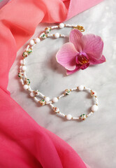 Necklace made of glass beads and couazonne beads on a background of pink chiffon drapery and orchid flower, composition with women's jewelry, selective focus, vertical orientation.