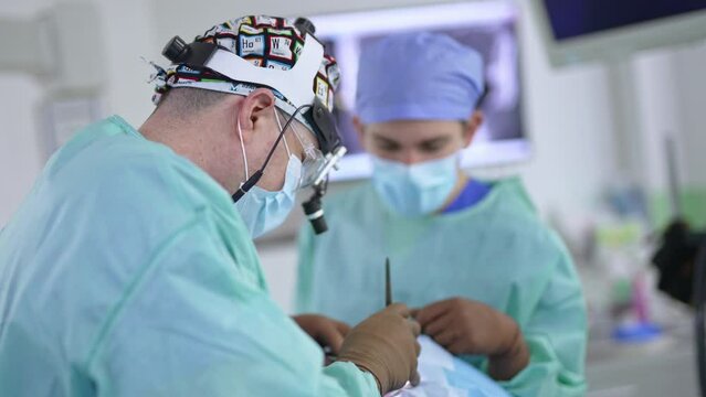 Male surgeon wearing device glasses, cap and mask performs surgery. Doctor standing his side to camera uses tool operating the patient.