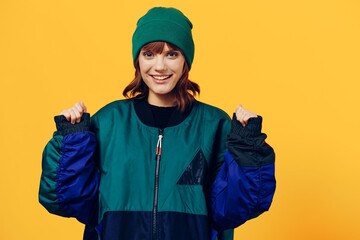 a cheerful woman in a green jacket and a hat in the style of the 90s stands on a yellow background happily looking at the camera and raises her hands up in a fist