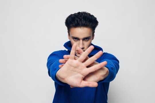 photo of a handsome, stylish man with thick black hair standing on a light gray background in a blue zip-up sweater and charmingly looking into empty space and covering the camera with his hand