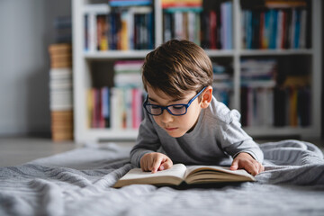 One caucasian boy lying on the floor at home in day reading a book front view wearing eyeglasses...