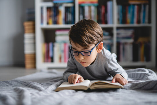 One caucasian boy lying on the floor at home in day reading a book front view wearing eyeglasses copy space real people