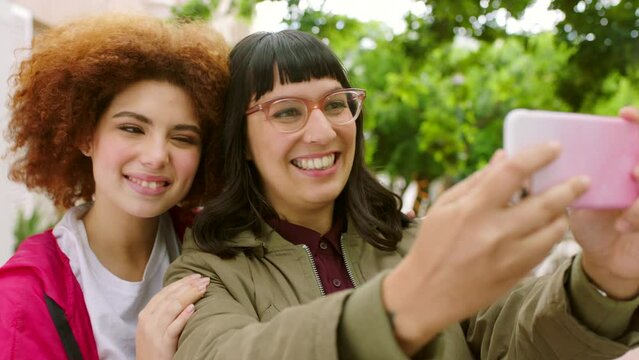Playful female friends taking selfies with a phone outside in an urban park. Happy girls smiling and posing for a photograph to post on social media. Young women laughing together making memories