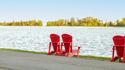 Two Red Adirondack Chairs on the green grass of a pier. Lookout on large blue lake with a perfect...