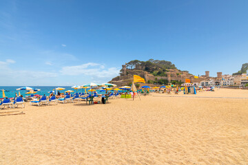 Spanish and tourists enjoy a sunny summer day at Platja Gran, the wide sandy beach at the Costa Brava town of Tossa de Mar with the 12th century Castle in view behind.