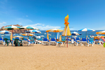 Beach chairs and colorful umbrellas along the sandy Platja Gran beach at the resort town of Tossa de Mar, Spain, on the Costa Brava coast of the Mediterranean Sea.