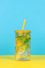 A large glass with water and lemon and a cocktail tube on a yellow and blue background.