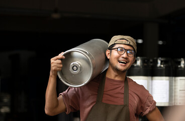 Young man in leather apron holding beer keg at modern brewery, craft brewery worker