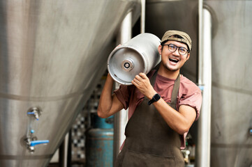 Young man in leather apron holding beer keg at modern brewery, craft brewery worker