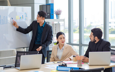 Businessman manager presenter in formal suit standing holding pen pointing at graph chart document on whiteboard presenting company information to Asian male female colleagues in meeting room