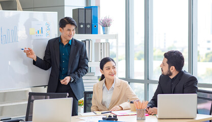 Businessman manager presenter in formal suit standing holding pen pointing at graph chart document on whiteboard presenting company information to Asian male female colleagues in meeting room