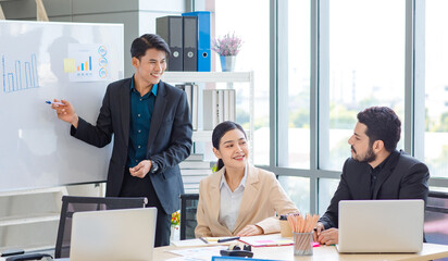 Businessman manager presenter in formal suit standing holding pen pointing at graph chart document on whiteboard presenting company information to Asian male female colleagues in meeting room