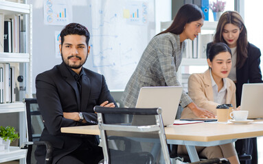 Millennial Asian Indian male confident businessman in formal suit sitting crossed arms looking outside when female businesswomen colleague working with laptop computer together in office meeting room