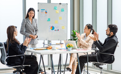 Fototapeta premium Asian businesswoman presenter in formal suit standing smiling holding pen pointing at graph chart document on whiteboard presenting information to multinational male female colleagues in meeting room
