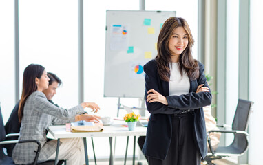 Portrait shot of millennial Asian cheerful successful professional businesswoman entrepreneur in formal suit standing smiling crossed arms in meeting room while employee and colleague brainstorming