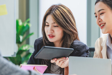 Closeup shot of two young millennial Asian professional cheerful female businesswomen in formal suit sitting holding tablet computer busy working with colleague in company conference meeting room