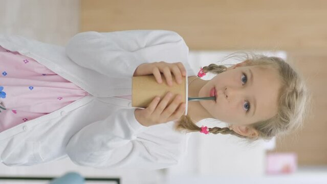 Vertical video. Cute little girl holding cardboard cup and drinking cacao or hot chocolate from a straw indoors