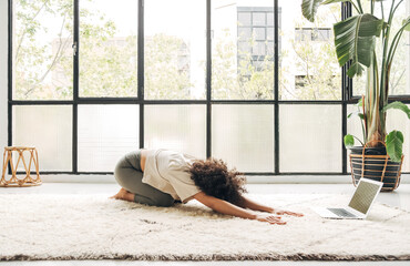 Young mixed race woman practicing yoga balasana pose at home following online classes using laptop....