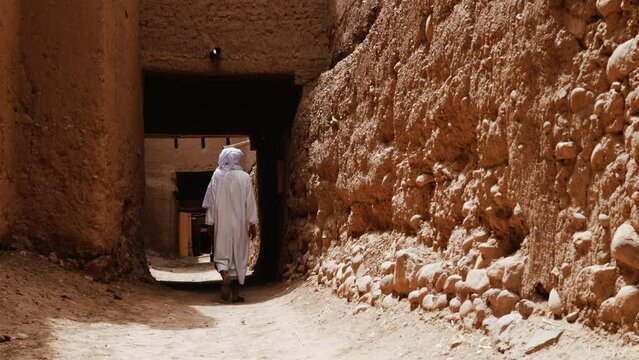 A traditional dressed Bedouin Moroccan man wearing a white gandoura and a turban walks inside a Kasbah in Tamnougalt, Morocco. Authentic rural scene of South Morocco. 4k.