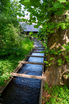 Vertical image of a colonial industry wooden sluice with flowing water in green nature setting going towards a historic industrial building in background