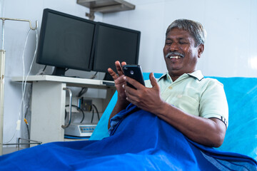 Happy smiling senior man using mobile phone at hospital while lying on bed - concept of medicare treatment, social media and relaxation