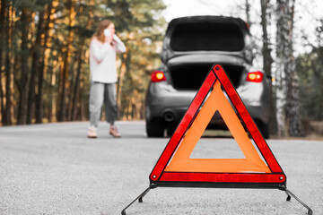 Red emergency triangle with blurred car and woman calling car mechanic in the background. Broken...