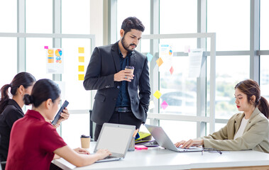 Millennial Indian bearded male businessman mentor standing holding disposable coffee cup teaching coaching Asian intern trainee secretary working with laptop computer in multinational company office