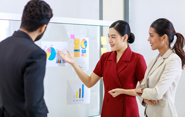 Fototapeta na wymiar Asian young beautiful professional successful businesswoman standing smiling holding hand presenting showing graph chart report data paperwork documents on glass board to male and female colleagues