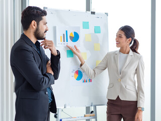 Asian businesswoman presenter in formal suit standing smiling holding pen pointing at graph chart document on whiteboard presenting information to multinational male female colleagues in meeting room.