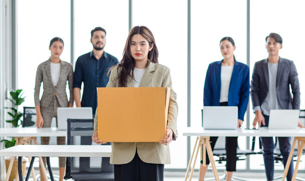Portrait shot of Asian sad jobless businesswoman in casual suit standing holding belongings in cardboard box after fired while male and female colleagues waving hands goodbye in blurred background
