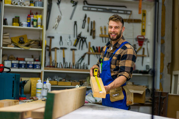 Portrait of young handsome caucasian carpenter who measures a board and smiling for camera