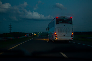 a view of a passenger white bus traveling along the highway on a night road at speed from the side of a car overtaking it at dusk in traffic