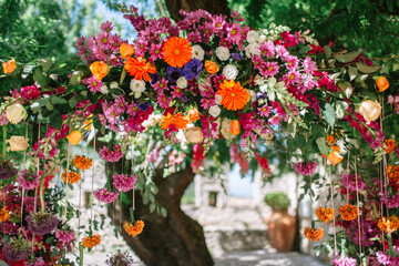 Italian floral arch with colorful flowers. Chrysanthemums, gerberas, roses, peonies, and hanging flowers tied with ribbons. In the background is a large tree.