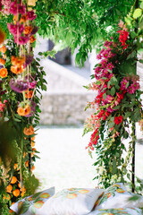 Fototapeta na wymiar An arch entwined with multi-colored bright flowers and vines in an Italian garden. In the background is a bench with many pillows.