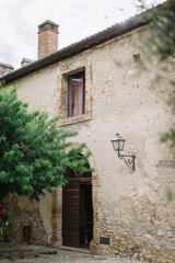 Old Italian stone house with an open arched wooden door, brick chimney, and wrought iron lantern. There is a big tree in front of the house.