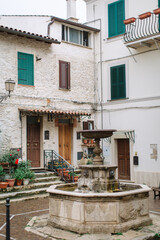 Italian patio with a classic stone fountain. In the background are white house facades with turquoise shutters and potted flowers on the steps.